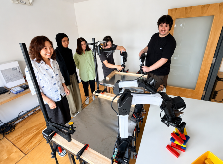 A group of people stand around a table. There are robotic arms on the table moving around colorful wooden blocks.